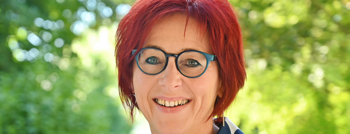 Frau mit kurzen roten Haaren und Brille im Porträt. Hintergrund grüne Sträucher.