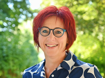 Frau mit kurzen roten Haaren und Brille im Porträt. Hintergrund grüne Sträucher.