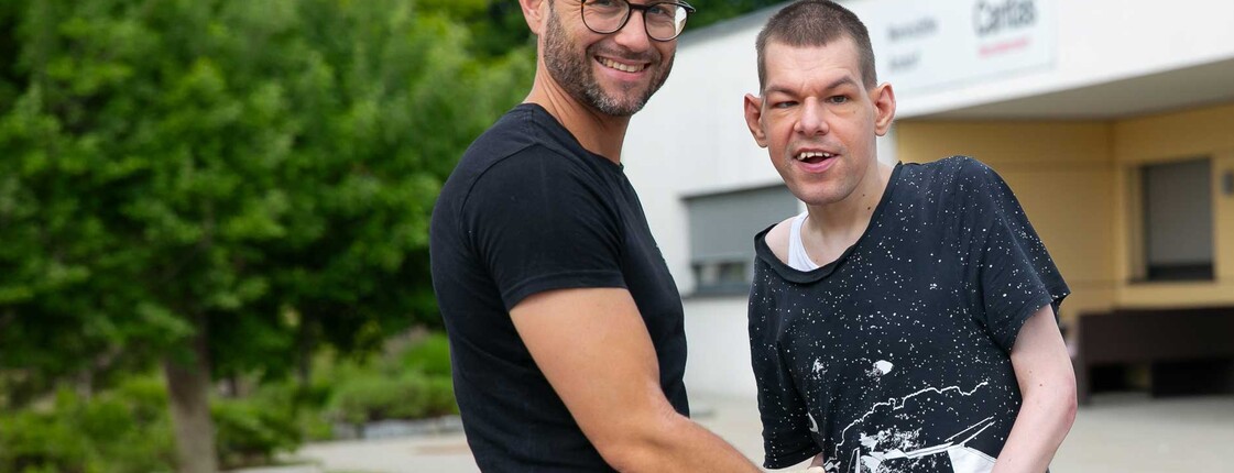 Zwei Männer stehen im Freien vor einem Caritas Gebäude und halten sich an den Händen.