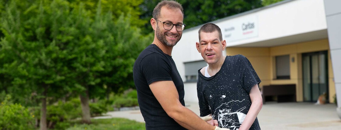 Zwei Männer stehen im Freien vor einem Caritas Gebäude und halten sich an den Händen.