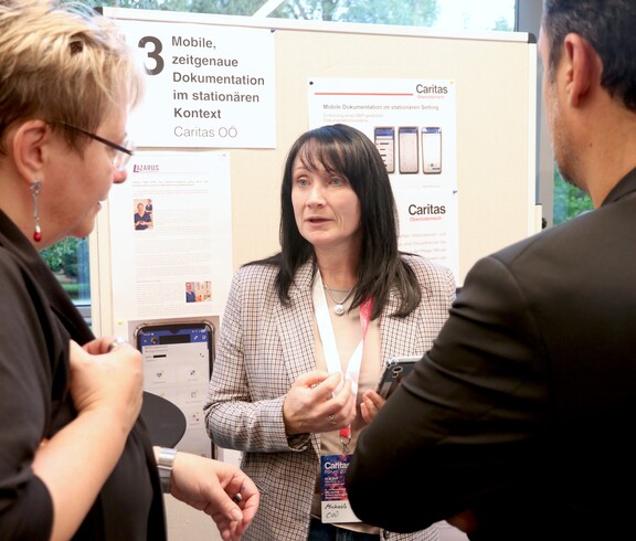 Eine Frau steht vor einer Pinnwan und erklärt zwei Personen etwas.