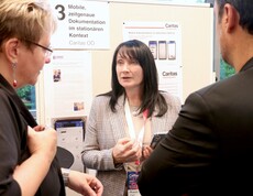 Eine Frau steht vor einer Pinnwan und erklärt zwei Personen etwas.