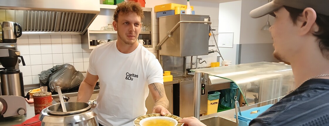 Mann im weißten T-Shirt reicht Mann im schwarzen T-Shirt einen gefüllten Suppenteller über die Theke.
