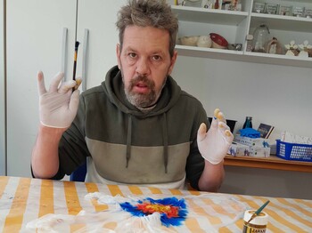 Mann hält seine Hände, die in Gummihandschuhen stecken in die Höhe, vor ihm liegt ein Stück bemalter Seidenstoff 