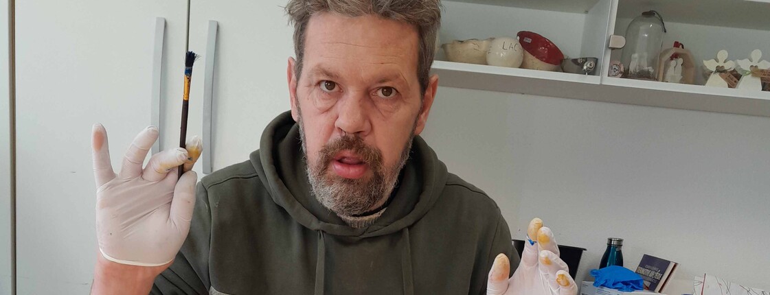 Mann hält seine Hände, die in Gummihandschuhen stecken in die Höhe, vor ihm liegt ein Stück bemalter Seidenstoff 