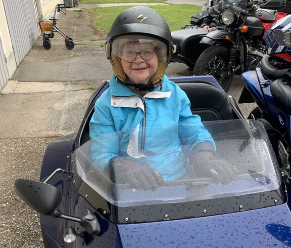 Ältere Frau mit Helm und Brille sitzt lächelnd im Beiwagen eines Motorrads vor dem Caritas-Gebäude
