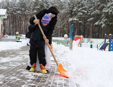 Eine Betreuerin hilft einem Kind beim Schneeschippen auf dem Winterspielplatz im Kinderdorf Gomel, umgeben von Spielgeräten und Schnee.