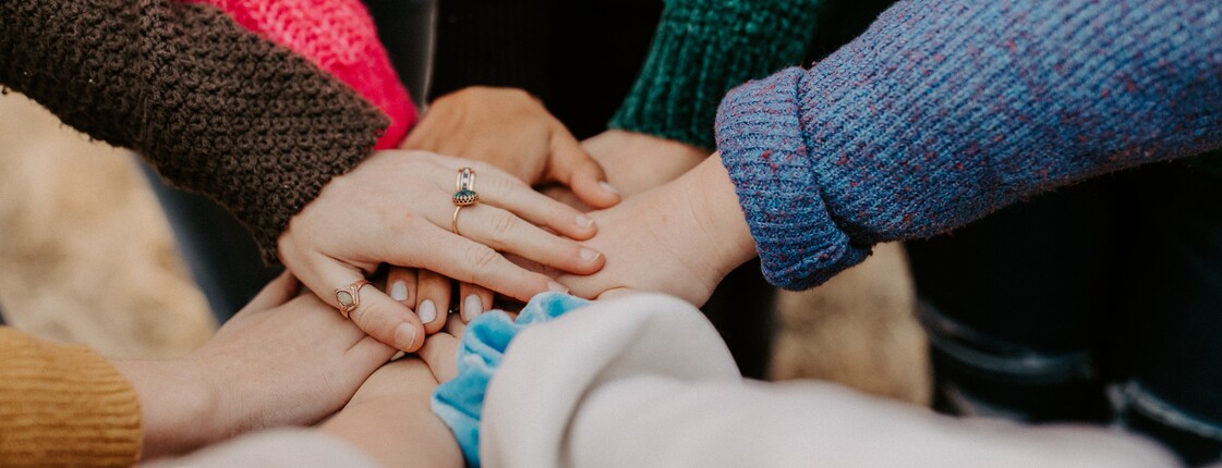 Gemeinschaft und Zusammenhalt – Caritas und Kirche Mehrere Hände unterschiedlicher Personen liegen übereinander als Zeichen von Gemeinschaft und Zusammenhalt. Die Personen tragen warme, bunte Strickpullover.