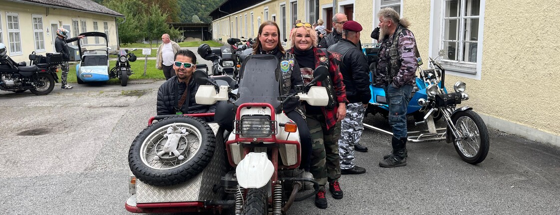 Drei Personen stehen bei einer Beiwagenmaschine. Im Hintergrund sind noch mehrer Motorräder zu sehen.