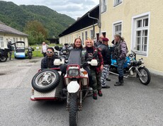 Drei Personen stehen bei einer Beiwagenmaschine. Im Hintergrund sind noch mehrer Motorräder zu sehen.