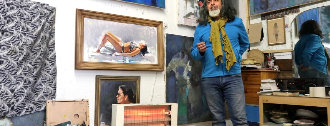 Ein Mann steht in seinem Atelier vor seinen Bildern, die an der Wand hängen.