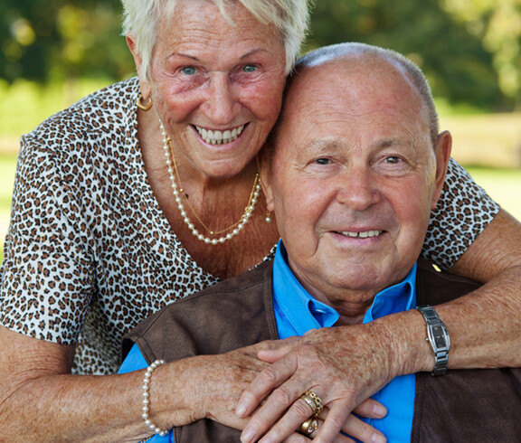 Ein Seniorenpärchen umarmt sich. Die Frau hat kurze blonde Haare, der Mann eine Glatze. Sie trägt eine leopardenfarbene Bluse, er ein blaues Hemd mit schwarzen Jackett. Sie wirken freundlich und enthusiastisch. 