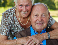 Ein Seniorenpärchen umarmt sich. Die Frau hat kurze blonde Haare, der Mann eine Glatze. Sie trägt eine leopardenfarbene Bluse, er ein blaues Hemd mit schwarzen Jackett. Sie wirken freundlich und enthusiastisch. 