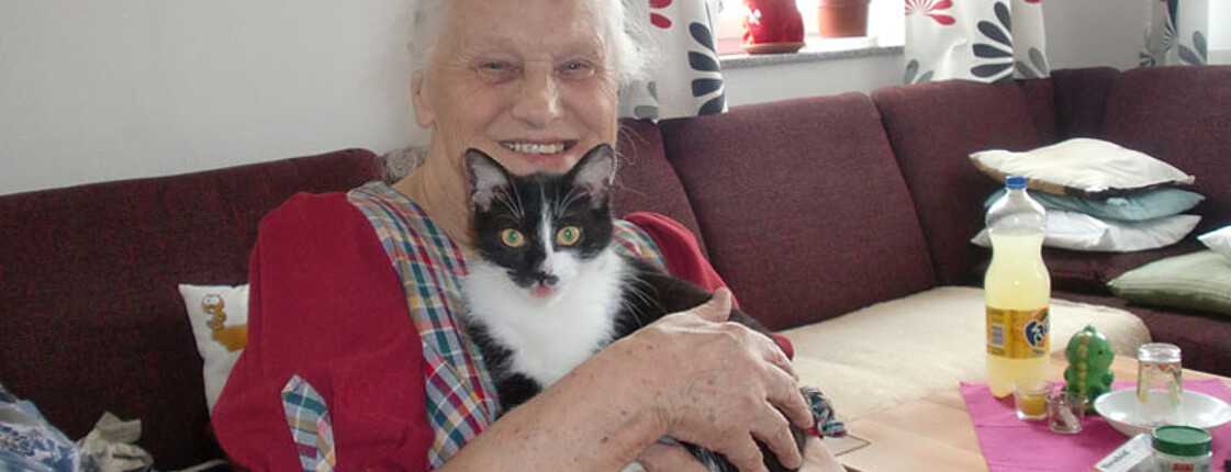 Eine ältere Frau sitzt in einem Wohnzimmer und hält eine Katze im Arm. 