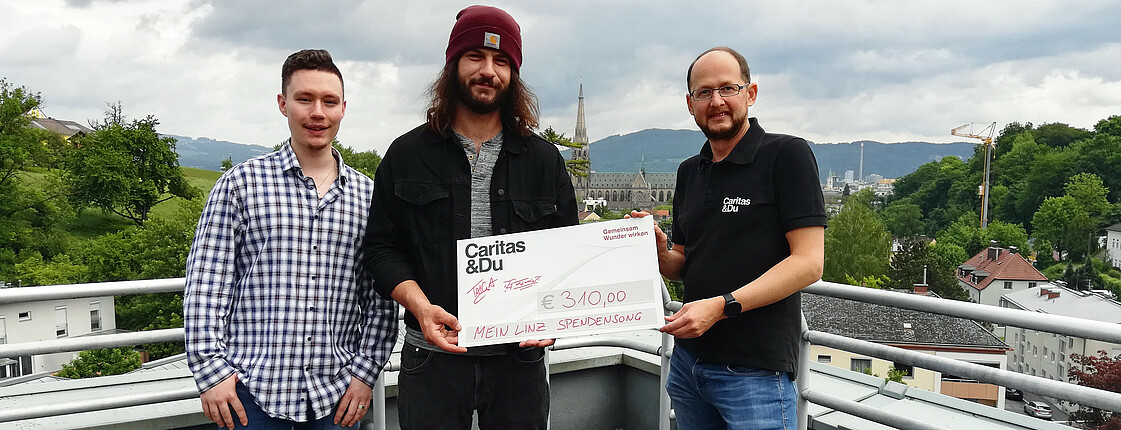 Zwei junge Männer der Musikgruppe „BluntKartell“ übergeben einen Spendencheck in Höhe von 310 Euro an Caritas-Mitarbeiter Andreas Falkner