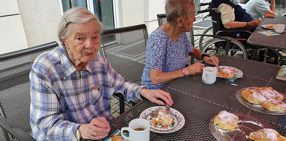 Zwei ältere Damen sitzen bei Tisch bei Kaffee und Kuchen. Dame links sieht ins Bild.