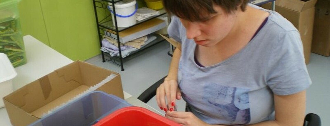 Eine junge sehbeeinträchtigte Frau sitzt an einem Arbeitstisch. Vor ihr ist eine Plastikbox mit kleinen Schachteln mit Tintenpatronen.