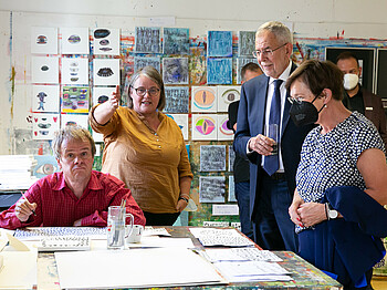 Vier Personen in der Kunstwerkstätte St. Pius. Rechts Bundespräsident Van der Bellen und seine Gattin, stehend neben einem Tisch mit Zeichungen.