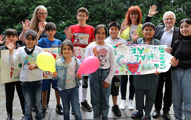 Gruppe stehender Kinder mit Luftballons und drei Erwachsene im Hintergrund.
