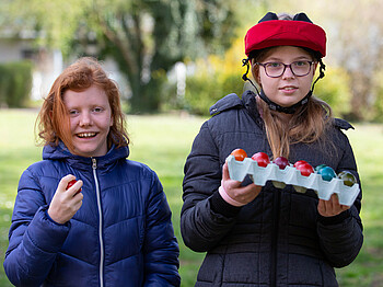 Zwei Mädchen halten auf einer Wiese gefärbte Ostereier in den Händen und freuen sich.