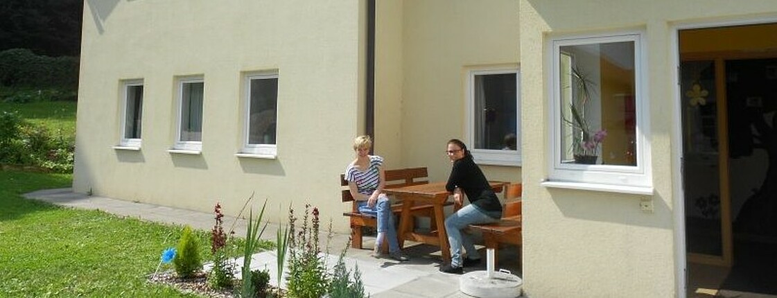 Blick auf ein Haus mit zwei Frauen, die auf einer Gartenbank sitzen.