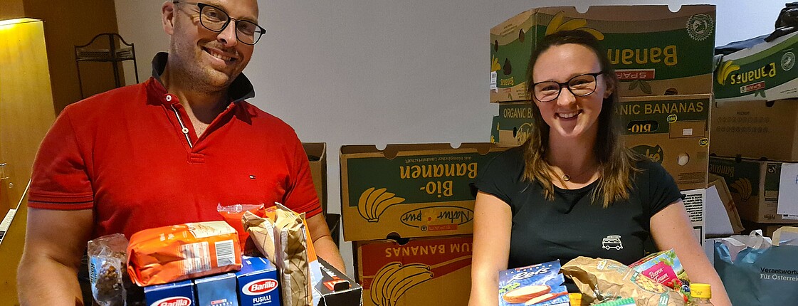 Zwei Menschen mit Kartons in der Hand, voll mit Lebensmitteln. Links ein Mann mit Brille, rechts eine junge Frau mit Brille und langen Haaren.
