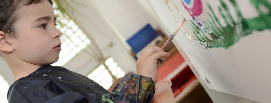 Ein kleiner Junge malt konzentriert mit einem Pinsel ein Bild auf ein großes Stück Papier.