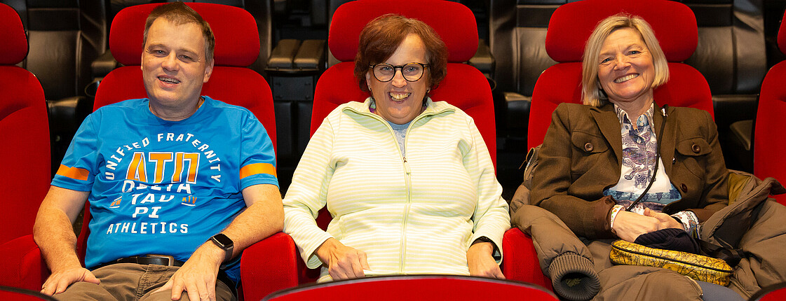 Drei Personen sitzen auf Kinosesseln und schauen in die Kamera