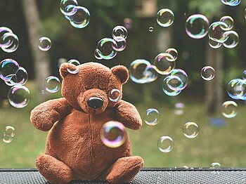 Brauner Teddybär sitzt mit geschlossenen Augen in Mitten von Seifenblasen.