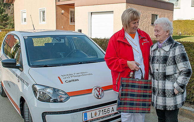 Eine Caritas-Mitarbeiterin steht mit einer älteren Frau vor dem Auto der Mobilen Dienste.