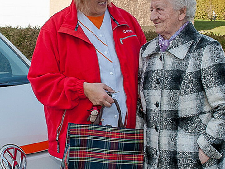 Eine Caritas-Mitarbeiterin steht mit einer älteren Frau vor dem Auto der Mobilen Dienste.