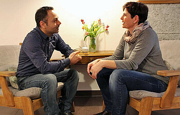 Eine Frau und ein Mann südländischer Herkunft sitzen auf gemütlichen Sesseln sich gegenüber und unterhalten sich.
