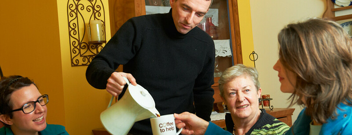 Mann steht in der Mitte und gießt sitzender Frau rechts Kaffee in eine Tasse. Zwei weitere Frauen sitzen im Hintergrund.