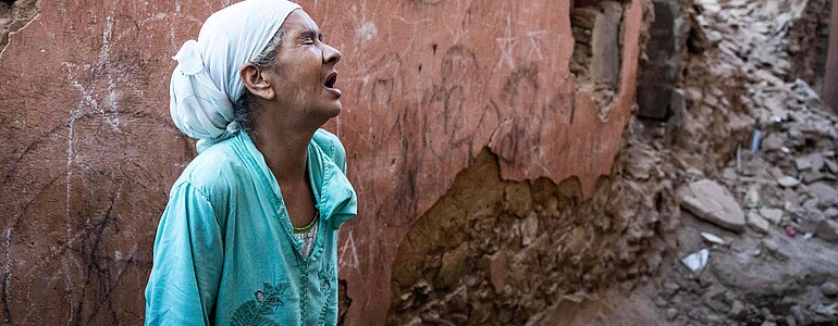 Eine vor Schmerz und Leid klagende Frau steht vor einer zerstörten Hauswand inmitten von Trümmern des Erdbebens. Sie hat einen weißen Turban und blaue Kleidung an.